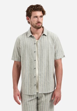 Mann trägt hellgraues kurzärmeliges Hemd mit Knöpfen und passende gestreifte Hose, schaut seitlich vor einfarbigem Hintergrund.