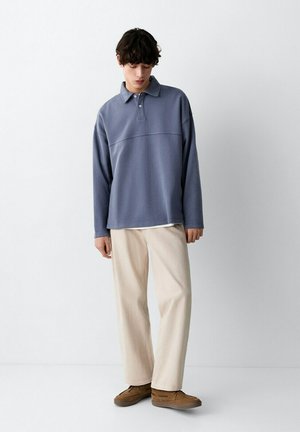 Joven con pantalones beige holgados, zapatos marrones y una camiseta polo azul de manga larga, de pie contra un fondo blanco liso.