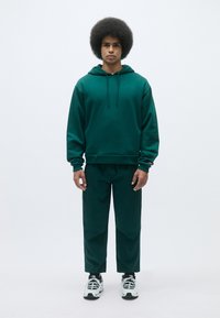 Felpa verde scuro e pantaloni abbinati realizzati in tessuto morbido. La felpa presenta un cordino e polsini a coste, con accenti in contrasto sulle maniche.