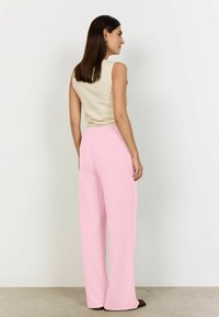 Mujer con cabello lacio y oscuro, vistiendo una blusa sin mangas color beige y pantalones anchos de tiro alto en color rosa, de pie de perfil contra una pared lisa.