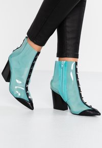 Bottes translucides turquoises à hauteur de cheville avec des pointes noires, des centres cloutés, des fermetures éclair sur les côtés, portées avec un pantalon noir ajusté sur fond blanc.