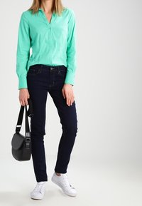 Camicia button-up verde menta con maniche lunghe, abbinata a jeans blu scuro. Sneakers bianche e una borsa a tracolla nera completano l'outfit.
