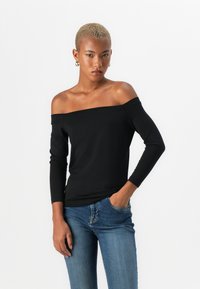 Top negro sin mangas con mangas largas, de tejido suave y diseño ajustado; combinado con jeans azules en una pose de pie frente a un fondo blanco.