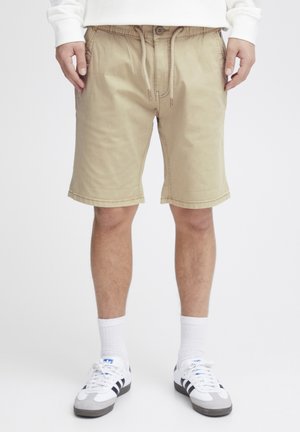 Beige Baumwollshorts mit elastischem Bund, Kordelzug, zwei seitlichen Taschen und gesticktem Saum. Getragen mit weißen Socken und Sneakers.