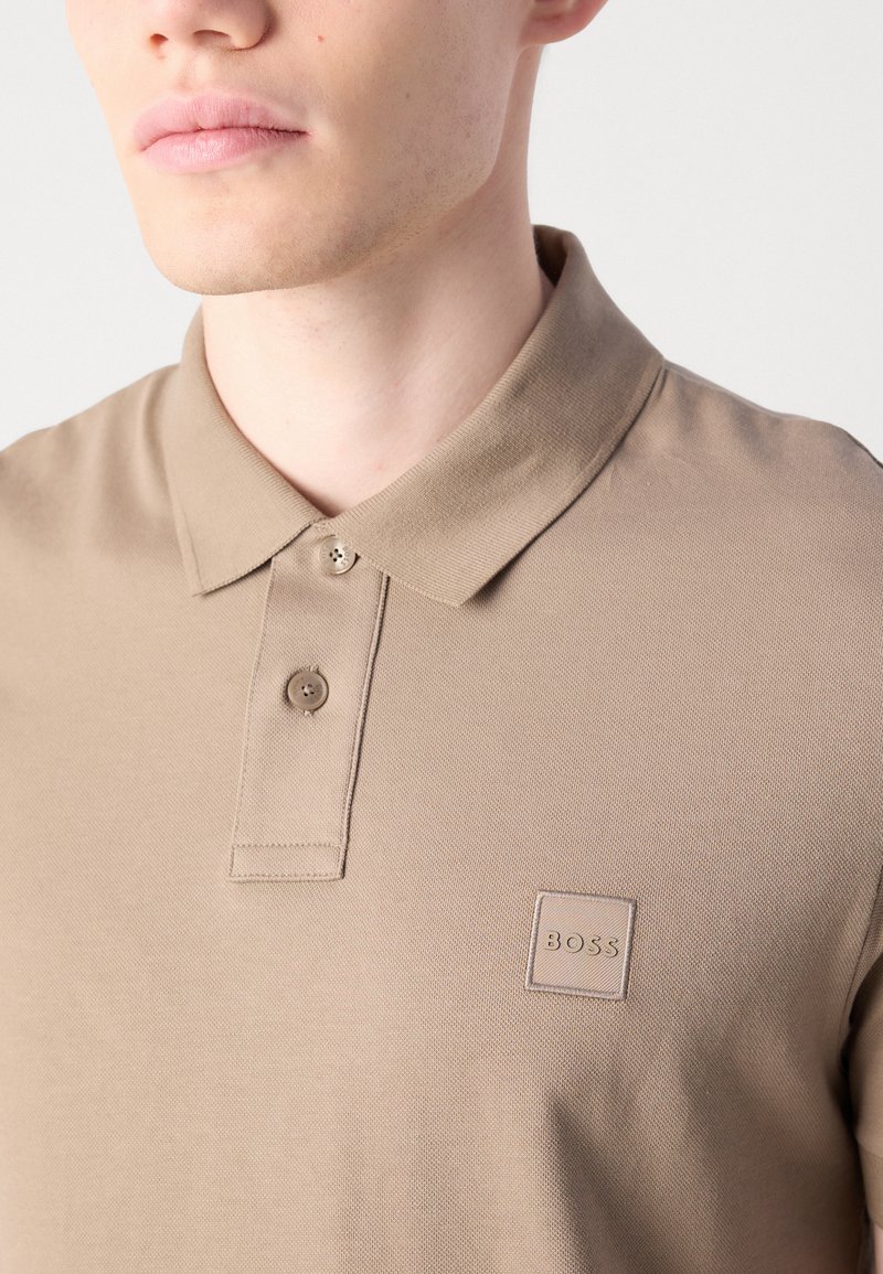 Gros plan sur une personne portant un polo beige avec une patte de boutonnage à deux boutons et un logo carré "BOSS" sur la poitrine gauche.