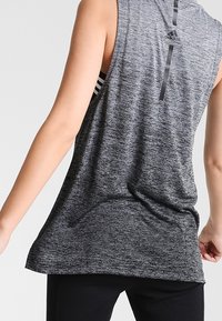 Harmaa hihaton urheilu-toppi, jossa on melange-tekstuuria, pyöristetty helma ja takana logo-yksityiskohta mustalla raidalla alla.