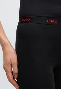 Leggings noirs avec une coupe ajustée, dotés d'une ceinture noire brodée du mot "HUGO" en rouge. Texture lisse, sans motifs ni accents.