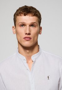 Vit linneskjorta med knappstängning och ståkrage, med en liten broderad design på bröstet. Slen textur, skräddarsydd passform.