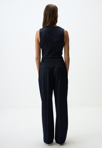 Marineblauwe mouwloze jumpsuit met een aansluitende bovenkant en wijde pijpen, voorzien van zakken en knoopaccenten op de achterkant van de taille. Glad stofgevoel.