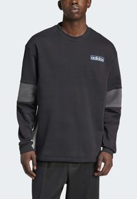 Černá mikina s kulatým výstřihem, dlouhými rukávy s šedými detaily a modrou nášivkou loga Adidas. Měkká látka, volný střih.