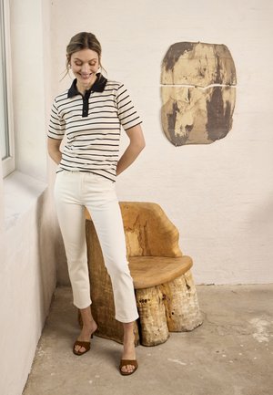 Femme en polo rayé et pantalon blanc debout près d'une chaise en bois rustique et d'une œuvre d'art murale abstraite dans une pièce minimaliste avec un sol en béton.