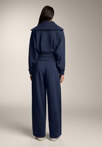 Nainen seisoo selin, hänellä on yllään tummansininen leveäkauluksinen collegepaita ja siihen sopivat leveälahkeiset housut sekä valkoiset kengät.