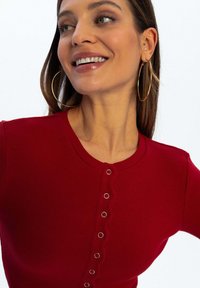 Czerwony sweter z prążkowanym wykończeniem i okrągłym dekoltem, z detalami w postaci guzików z przodu. Posiada subtelny teksturę i dopasowany krój. Modelka nosi duże kolczyki w kształcie kół.