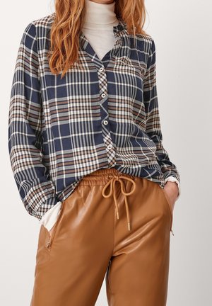 Kvinde iført en skjorte med navyblåt, brunt og hvidt ternet mønster over en hvid rullekrave, kombineret med brune læderbukser med snøre i taljen og hænderne i lommerne.