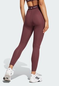Leggings bordeaux avec une taille haute, texture lisse, agrémentés d'un logo sur la taille, associés à des baskets blanches.