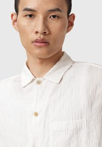 Chemise blanche en lin à boutons, avec une finition texturée, un col pointu, une poche poitrine et un bouton simple au niveau du col.