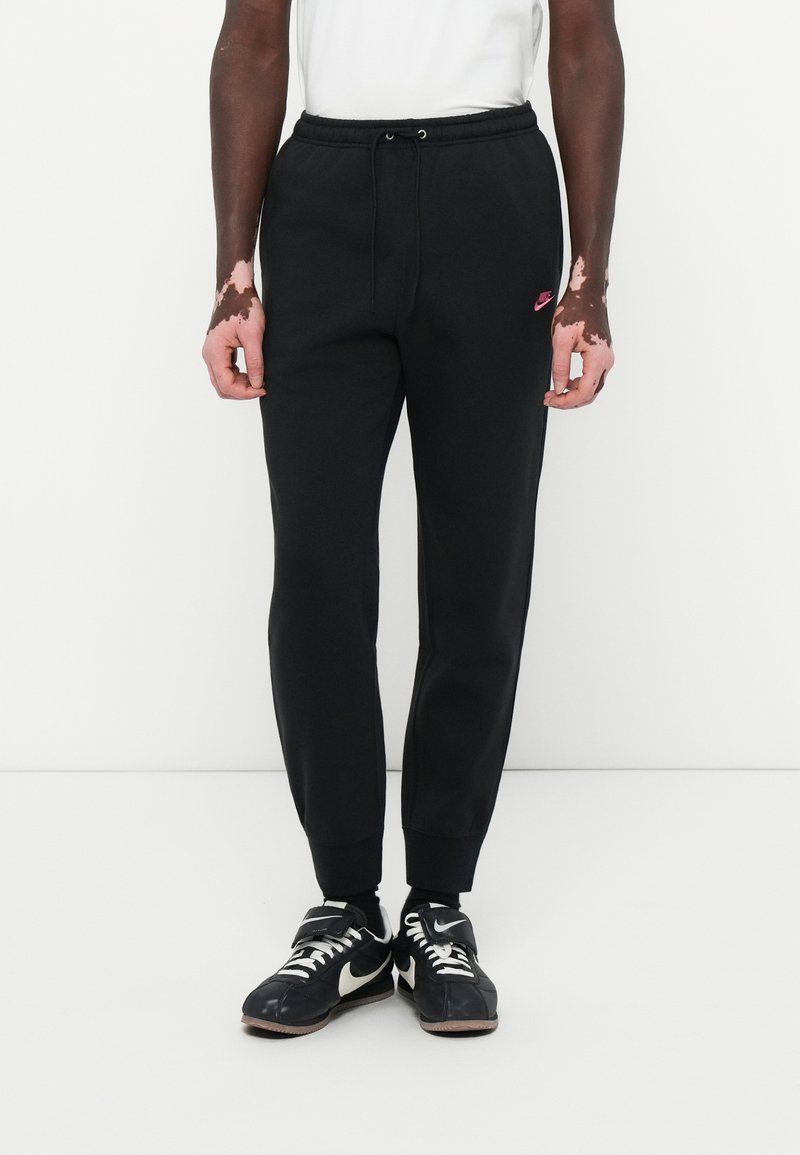 Sorte sweatpants med snørretalje, ribbede ankler og et lille pink Nike-logo på venstre ben. Båret med sorte Nike sneakers.