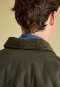 Kurtka z pikowanej oliwkowej tkaniny z prążkowanym kołnierzem, z niebieską bawełnianą warstwą widoczną przy dekolcie, gładka w dotyku, z ukośnym wzorem szycia.