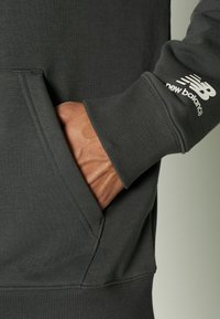 Sudadera gris oscuro con bolsillo tipo canguro, puños acanalados y el logo blanco de "New Balance" en la manga. Textura suave de mezcla de algodón.