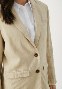 Mujer vistiendo un blazer de lino beige con botones marrones sobre una camisa blanca con cuello, de pie contra un fondo liso.