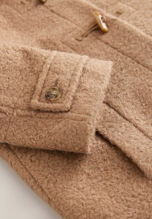 Hihainen vaaleanruskea villatakki, jossa on teksturoidu pinta, napitus ja läpällinen tasku, korostaen pehmeää, pörröistä materiaalia.