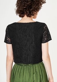 Top corto in pizzo nero con maniche corte e motivo floreale, abbinato a una gonna plissettata verde. Il design è aderente e texture.
