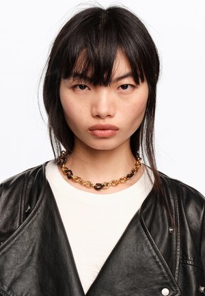 Joven con cabello negro liso, lleva un collar de cadena dorado y negro, blusa blanca y chaqueta de cuero negra, sobre un fondo blanco liso.