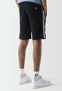 Pantalones cortos deportivos negros de material suave con rayas laterales. Presenta un bolsillo trasero y un pequeño logo en la cintura. Combinados con zapatillas blancas.