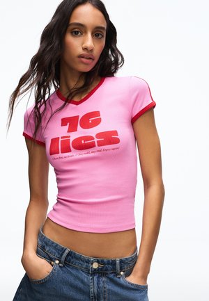 Žena s dlhými tmavými vlasmi, ktorá má na sebe ružové priliehavé tričko s červeným lemovaním a červeným nápisom, doplnené modrými rifľami, pózuje s rukami v vreckách.