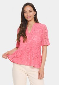 Rožinė nėriniuota palaidinė su trumpomis rankovėmis ir V formos iškirpte. Papuošta gėlėtų raštų ir turi peplumo apatinę dalį, demonstruojama kartu su šviesios spalvos kelnėmis.