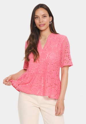 Top in pizzo rosa con maniche corte e scollo a V. Presenta motivi floreali e un orlo a peplum, mostrato sopra pantaloni chiari.