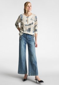 Blusa beige chiaro con stampa di navi blu e scollatura a chiave, abbinata a jeans in denim sbiadito a gamba larga con dettagli a bottone. Tacchi neri.