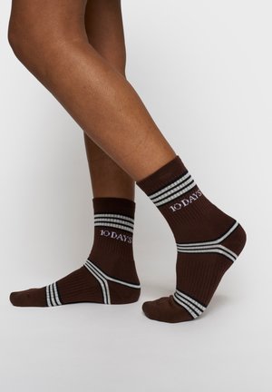 Gambe con calzini alla caviglia marroni con strisce bianche e nere e la scritta "10 DAYS" sul lato, un piede appoggiato a terra, l'altro leggermente sollevato.