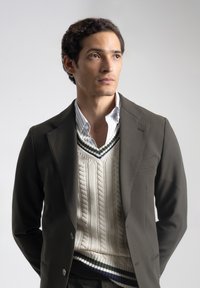 Joven con cabello rizado y oscuro, vestido con una chaqueta oscura sobre un suéter blanco de punto de cable y una camisa a rayas, mirando ligeramente hacia arriba y a la derecha.