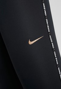 Czarne legginsy sportowe z fakturowanym wykończeniem, z złotym logo Nike i białą taśmą z nadrukiem wzdłuż boku.