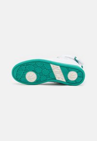 Semelle d'une basket Nike blanche et turquoise montrant une adhérence texturée, logos de basket-ball sur le talon et la zone des orteils, et marquage Nike sur la semelle intermédiaire.