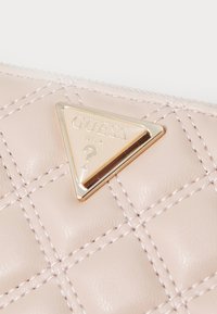 Portafoglio in pelle trapuntata rosa con un accento triangolare in metallo dorato del logo Guess, con testo inciso e un sottile dettaglio di cucitura.