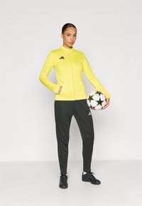 Κίτρινο μπουφάν με φερμουάρ και δύο τσέπες, μαύρες λεπτομέρειες και λογότυπο· σκούρα παντελόνια τζόγκινγκ και μαύρα αθλητικά παπούτσια· κρατώντας μια ποδοσφαιρική μπάλα με σχέδια.