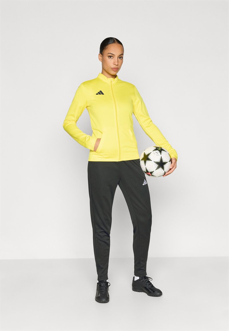 Κίτρινο μπουφάν με φερμουάρ και δύο τσέπες, μαύρες λεπτομέρειες και λογότυπο· σκούρα παντελόνια τζόγκινγκ και μαύρα αθλητικά παπούτσια· κρατώντας μια ποδοσφαιρική μπάλα με σχέδια.