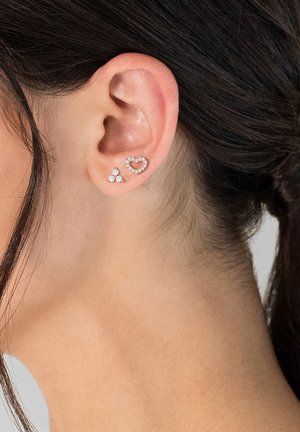 Două cercei expuși pe o ureche: unul în formă de inimă și celălalt triunghiular, amândouă cu pietre de zirconiu cubic clare și un finisaj metalic.