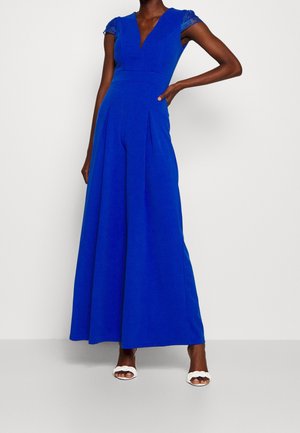 Frau trägt einen königsblauen Jumpsuit mit weitem Bein und Spitzen-Kappenärmeln sowie einem tiefen V-Ausschnitt, kombiniert mit weißen Sandalen mit geflochtenen Riemen und Absatz.