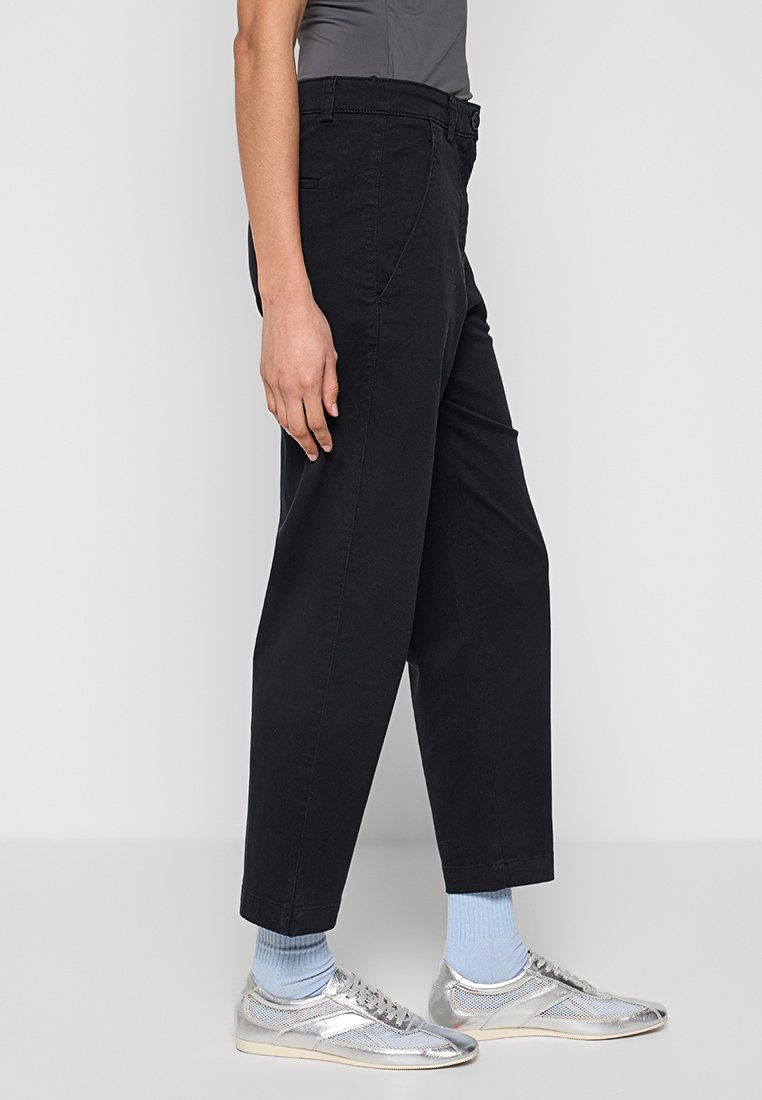 Schwarze Baumwollhosen mit geradem Bein, ausgestattet mit seitlichen Taschen und einem mittelhohen Bund. Getragen mit silbernen Sneakers und hellblauen Socken.