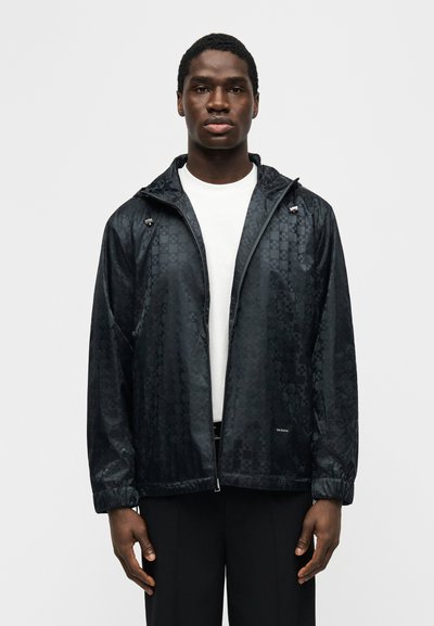 Άνδρας μοντέλο που φοράει μαύρο μοτίβο ζακέτας με φερμουάρ πάνω από άσπρο πουκάμισο, στέκεται μπροστά σε απλό φωτεινό φόντο, κοιτώντας μπροστά.