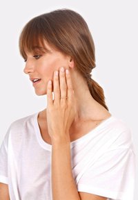 Anello d'oro su una mano che accarezza la guancia, indossato con una camicia bianca casual. I capelli sono castani e acconciati con onde morbide, con una riga laterale.