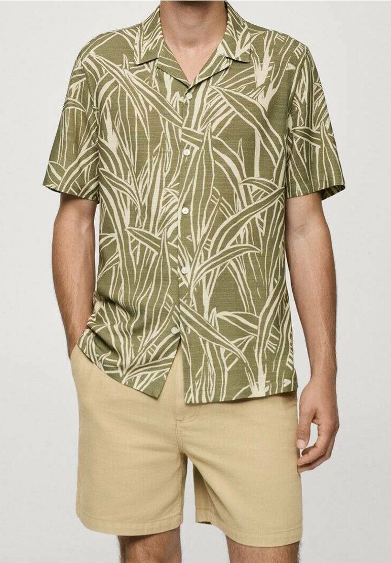 Homme portant une chemise boutonnée à manches courtes vert olive avec motif de feuilles beige et un short beige, une main dans la poche, sur fond uni.