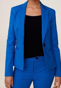 Mujer vestida con un brillante blazer azul y pantalones a juego con una blusa negra, de pie contra un fondo neutral.