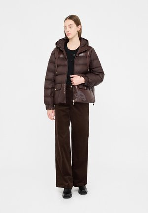 Giacca imbottita marrone con finitura lucida, dotata di cappuccio, tasche frontali e accessori in tono oro, indossata sopra una maglia nera e pantaloni larghi marroni.