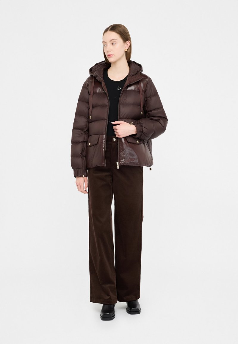 Giacca imbottita marrone con finitura lucida, dotata di cappuccio, tasche frontali e accessori in tono oro, indossata sopra una maglia nera e pantaloni larghi marroni.