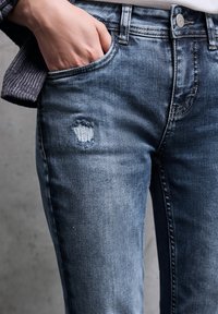 Haalarin siniset farkut, joissa on kulunut design, jossa vasemmassa reidessä on selkeä reikä ja viisi taskua.
