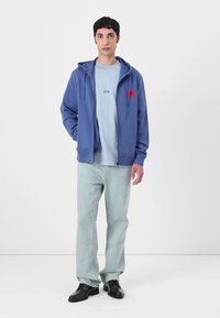 Blå zip-up hettegenser med et rødt logo-lapp, lagd over en lys blå t-skjorte, kombinert med lyse jeans og svarte sko. Jevn stofftekstur.